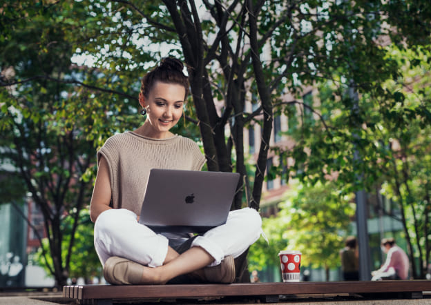 Женщина сидит на лавочке под деревом и работает в ноутбуке